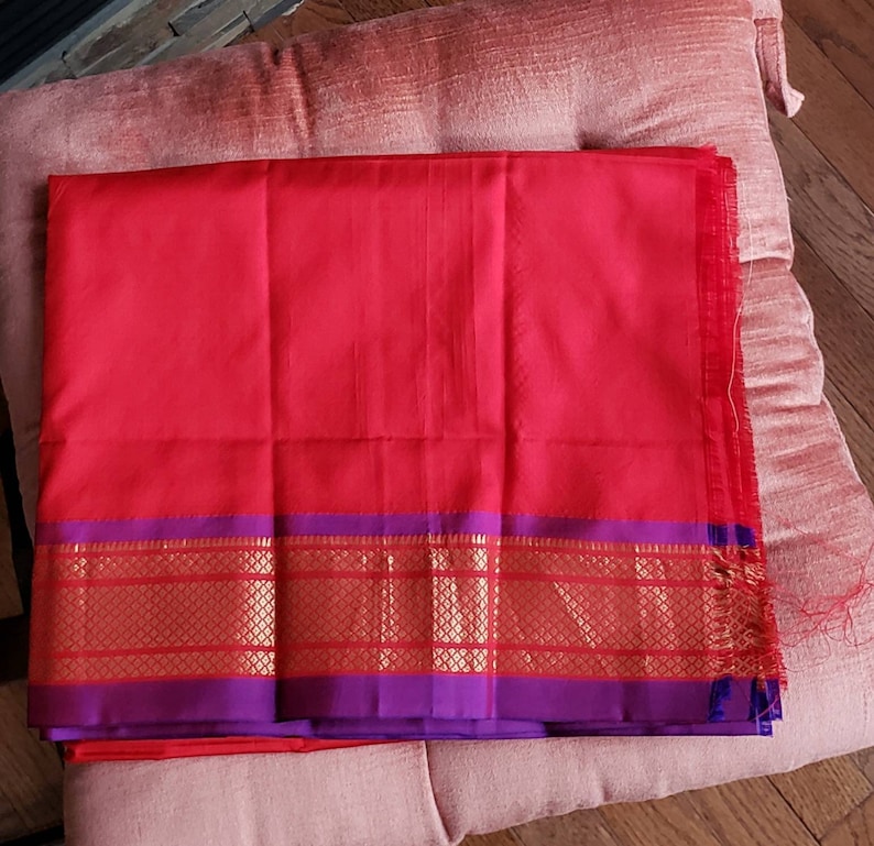 May include: A red and purple silk saree with a gold border. The saree is folded and lying on a pink cushion.