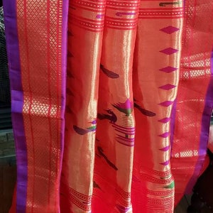 May include: A red and gold saree with a purple border. The saree has a traditional Indian design with intricate patterns and a peacock motif.