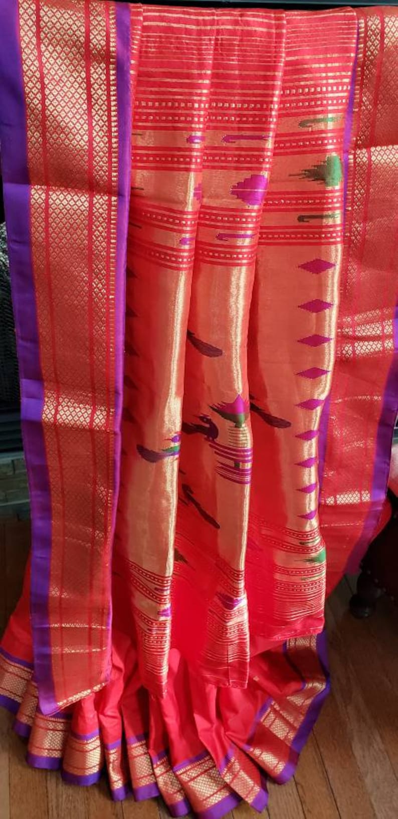 May include: A red and gold saree with a purple border. The saree has a traditional Indian design with intricate patterns and a peacock motif.