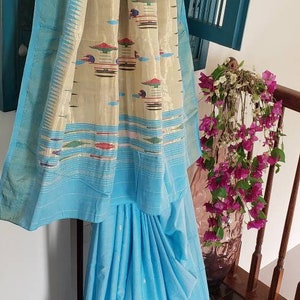 May include: A light blue saree with a cream and multi-colored patterned border. The saree is draped over a hanger and has a light blue pallu with white dots.