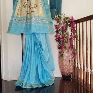 May include: A light blue saree with a gold and white patterned border. The saree is draped over a hanger and is hanging in front of a blue wooden mirror. There are pink flowers in a vase to the right of the saree.