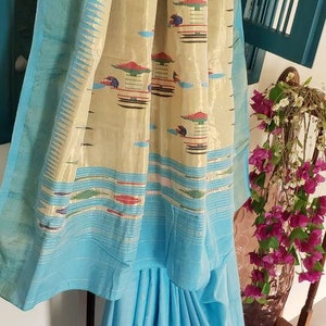 May include: A light blue saree with a cream and gold border. The border has a repeating pattern of colorful geometric shapes and designs. The saree is draped over a wooden stand.