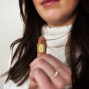 May include: A gold chain necklace with a square pendant featuring the letter "V" in a raised design. The pendant has a textured border.