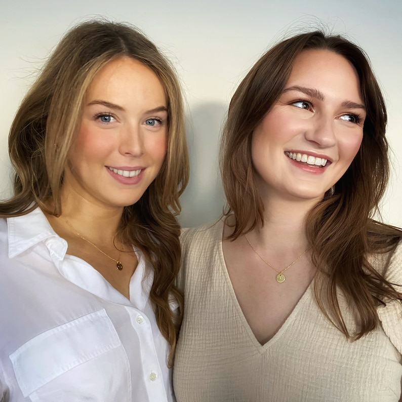 May include: Two women with long brown hair are smiling at the camera. They are both wearing gold necklaces with small round pendants.