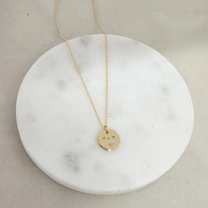 May include: A gold necklace with a round pendant that has the letters "ALS" engraved on it. The pendant is on a white marble surface.