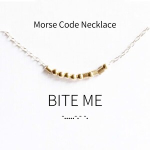 May include: A gold necklace with a silver chain and gold beads spelling out "BITE ME" in Morse code. The necklace is on a white background with the phrase "Morse Code Necklace" and the Morse code for "BITE ME" written below.