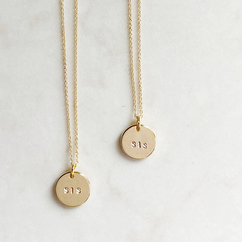 May include: Two gold necklaces with round pendants that say "SIS" on them. The necklaces are on a white background.