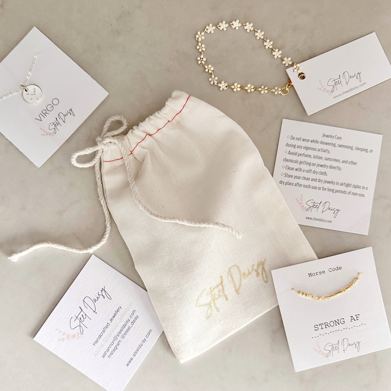 May include: A set of three gold necklaces with a white daisy chain, a Virgo zodiac sign charm, and a Morse code charm that reads "STRONG AF". The necklaces are packaged in a white drawstring bag with the brand name "Steel Daisy" printed on it. The image also includes two cards with the brand name and care instructions.