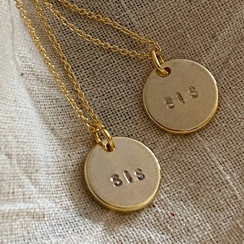 May include: Two gold necklaces with round pendants that say "SIS" on them. The necklaces are on a beige fabric background.