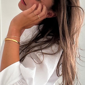 May include: A set of gold-toned jewellery including two rings worn on the fingers and three stacked bracelets on the wrist. The bracelets consist of a thin beaded chain, a smooth solid bangle, and a delicate plain chain, all in a polished gold finish.