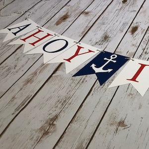 Puede incluir: Una pancarta de Ahoy It's a Boy con letras azules y rojas y anclas sobre un fondo blanco. La pancarta est&aacute; colgada sobre un fondo de madera blanqueada.