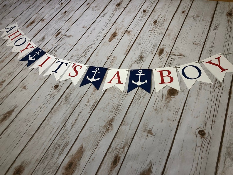 Puede incluir: Una pancarta de Ahoy It's A Boy con banderines rojos, blancos y azules. Cada bander&iacute;n tiene una letra o un s&iacute;mbolo de ancla diferente. La pancarta est&aacute; enhebrada en una cuerda blanca.