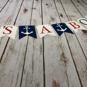 Puede incluir: Una pancarta blanca, roja y azul con el texto "It's a Boy" y anclas. La pancarta est&aacute; colgada en una superficie de madera.