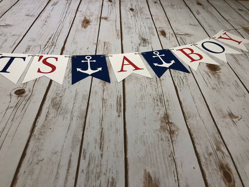 Puede incluir: Una pancarta blanca, roja y azul con el texto "It's a Boy" y anclas. La pancarta est&aacute; colgada en una superficie de madera.