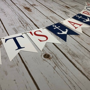 Puede incluir: Una pancarta blanca con letras rojas y azules que dice "It's a Boy" con un s&iacute;mbolo de ancla en las banderas "A" y "Boy". La pancarta est&aacute; atada con un cordel y est&aacute; colgada en una superficie de madera.