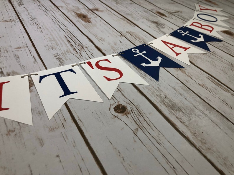 Puede incluir: Una pancarta blanca con letras rojas y azules que dice "It's a Boy" con un s&iacute;mbolo de ancla en las banderas "A" y "Boy". La pancarta est&aacute; atada con un cordel y est&aacute; colgada en una superficie de madera.