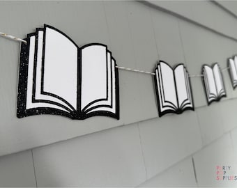 May include: A black and white book-shaped banner with glitter accents hangs on a gray wall. The banner is made of paper and is strung together with white string.