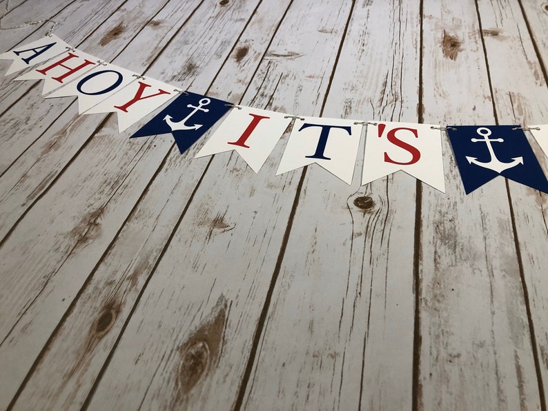 Puede incluir: Una pancarta de Ahoy It's a Boy con letras azules y rojas y anclas sobre un fondo blanco. La pancarta est&aacute; colgada sobre un fondo de madera blanqueada.