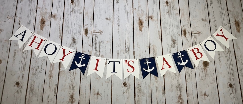 Puede incluir: Una pancarta de Ahoy It's a Boy con letras rojas y azules y anclas en banderines blancos. La pancarta est&aacute; enhebrada en un fondo blanco.