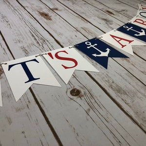 Puede incluir: Una pancarta blanca y azul con letras rojas que dice "It's a Boy" con un s&iacute;mbolo de ancla entre las palabras "a" y "boy".