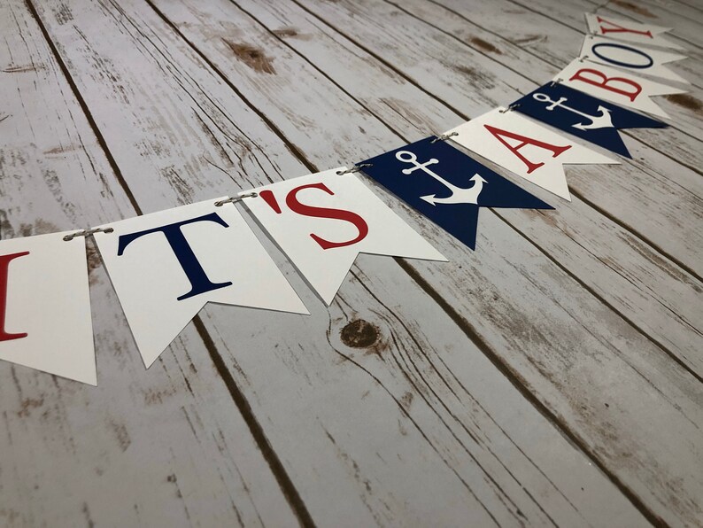 Puede incluir: Una pancarta blanca y azul con letras rojas que dice "It's a Boy" con un s&iacute;mbolo de ancla entre las palabras "a" y "boy".