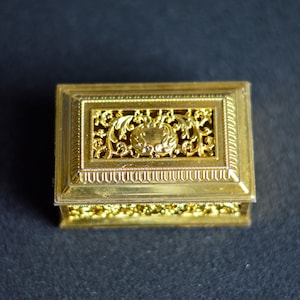 Può includere: Una piccola scatola rettangolare dorata con un design intricato e ornato. La scatola ha un bordo decorativo rialzato e un motivo floreale sul coperchio.