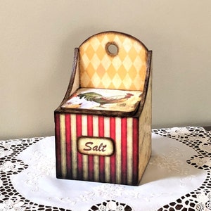 Könnte beinhalten: Ein hölzerner Salzstreuer mit rot-weißem Streifenmuster und einer Hühnerillustration. Der Streuer hat ein gelbes und weißes Rautenmuster auf der Oberseite und das Wort "Salz" auf der Vorderseite.