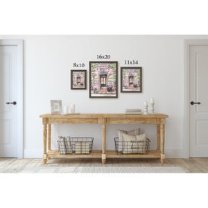 May include: Three framed prints of a pink storefront with green vines and a sign that reads "Boutique". The prints are displayed on a wall above a wooden console table with two black wire baskets. The prints are labeled with their dimensions: 8x10, 16x20, and 11x14.