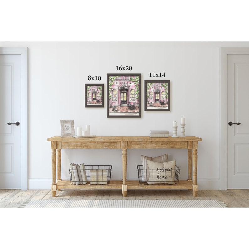May include: Three framed prints of a pink storefront with green vines and a sign that reads "Boutique". The prints are displayed on a wall above a wooden console table with two black wire baskets. The prints are labeled with their dimensions: 8x10, 16x20, and 11x14.