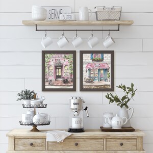 May include: Two framed watercolor paintings of Parisian-style shops with pink and blue awnings, hanging above a wooden shelf with white mugs and a white pitcher. Below the shelf is a wooden countertop with a coffee maker, a tiered stand with white mugs, and a white pitcher with a teacup.