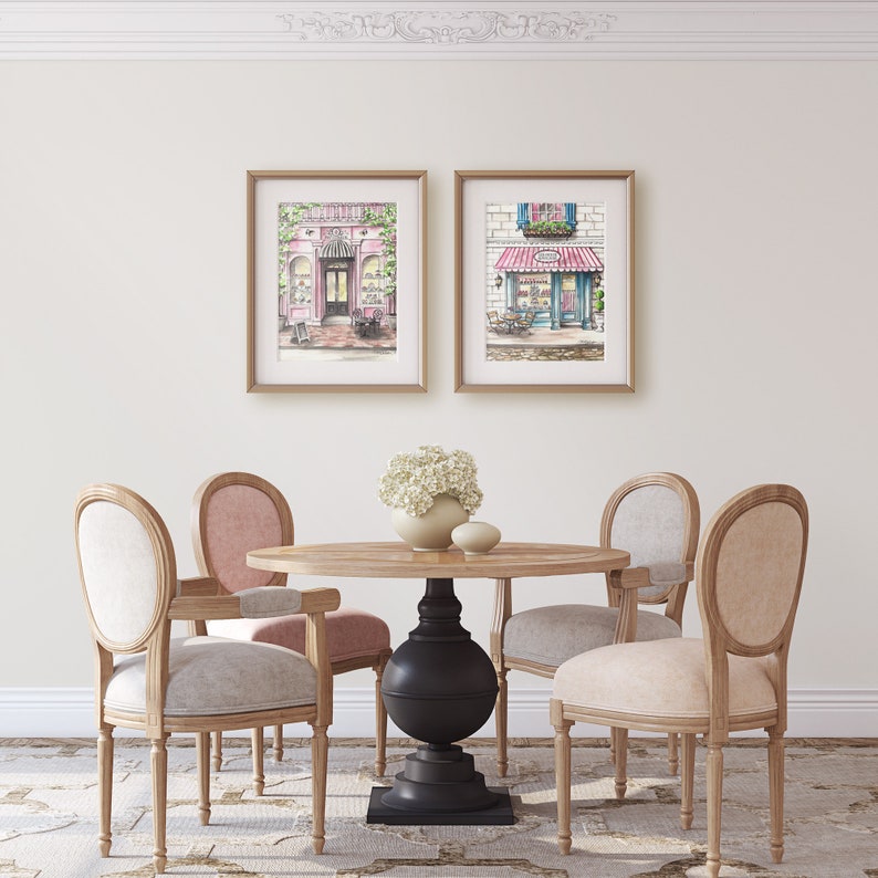 May include: Two framed illustrations of Parisian storefronts, one pink and one blue, hanging on a white wall above a wooden dining table with four chairs. The table has a black pedestal base and a light wood top. The chairs are light wood with beige and pink upholstery.