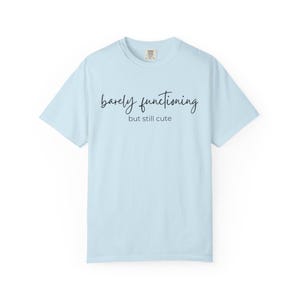 Puede incluir: Camiseta azul claro con la frase "barely functioning" en cursiva, sobre la frase "but still cute" en una fuente más pequeña y sin serifa. La camiseta tiene cuello redondo y mangas cortas.