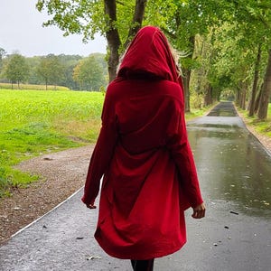 May include: A person wearing a long, hooded red coat walking on a wet, paved path. The coat is the focal point, with its vibrant color contrasting against the green landscape and overcast sky. Black leggings and boots complete the outfit.