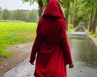 Op de afbeelding: Een persoon draagt een lange, rode jas met capuchon en loopt op een nat, geplaveid pad. De jas is het middelpunt, met zijn levendige kleur die contrasteert met het groene landschap en de bewolkte lucht. Zwarte leggings en laarzen maken de outfit compleet.