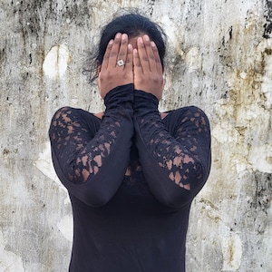 May include: Black long-sleeved top with lace sleeves and black pants with fringe detailing. The person is wearing a ring. The background is a weathered, textured wall.
