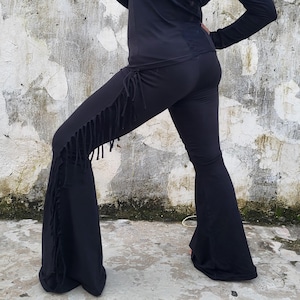 May include: Black long-sleeve top with lace detailing on the back, paired with black flared pants. The pants feature fringe detailing along the sides, creating a bohemian style. The outfit is made of a soft, flowing fabric.