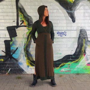 Puede incluir: Un vestido largo de color verde oscuro con capucha, cinturón marrón y aberturas laterales. El vestido lo lleva puesto una persona de pie frente a una pared de ladrillo cubierta de grafitis.