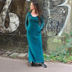 Puede incluir: Un vestido maxi de color turquesa con capucha y bolsillos. El vestido está hecho de una tela suave y texturizada y tiene un corte holgado. La modelo está de pie frente a una pared cubierta de grafitis.