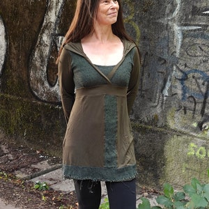 Może przedstawiać: Długa tunika z kapturem i wzorem patchworkowym w kolorze zielonym i brązowym. Tunika ma okrągły dekolt i luźny krój.