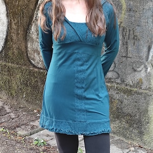 Puede incluir: Vestido de manga larga color turquesa con escote cuadrado y cintura entallada. El vestido presenta detalles de encaje en el corpiño y el dobladillo. La modelo lleva leggings negros. El fondo es una pared de hormigón desgastado.