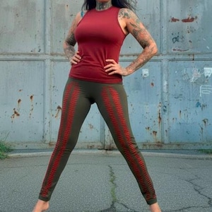 Puede incluir: Una camiseta sin mangas burdeos y leggings verde oliva con rayas rojas. La ropa deportiva la lleva una persona tatuada, posando frente a una puerta industrial roja y gris.