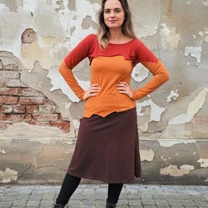 Könnte beinhalten: Eine Frau trägt ein mehrschichtiges langärmliges Oberteil in Orange- und Rottönen, kombiniert mit einem braunen Rock und schwarzen Leggings. Sie trägt auch schwarze Stiefel. Der Hintergrund ist eine verwitterte Wand.