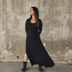 May include: A long black dress with a hood and long sleeves. The dress has a slit on the side and is paired with black boots. The person is standing in front of a textured gray wall.