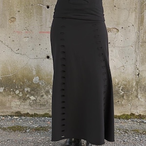 Może przedstawiać: Długa, czarna spódnica z wysokim stanem i serią małych, równomiernie rozmieszczonych wycięć biegnących pionowo wzdłuż boków. Spódnica wykonana jest z gładkiej, nieprzezroczystego materiału i sięga do kostek.