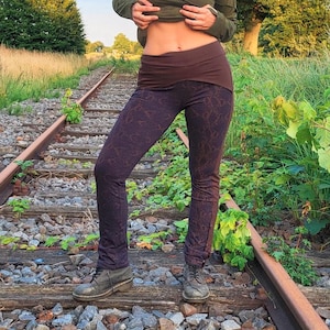 May include: Dark purple leggings with a brown waistband and a subtle snake-skin pattern. The leggings are paired with a long-sleeved olive green top. The model is standing on a railroad track.