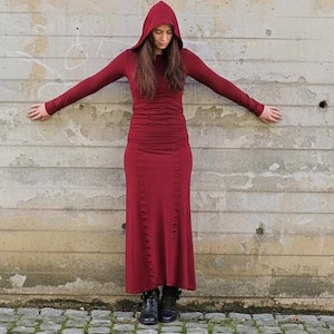May include: A long, burgundy hooded dress with long sleeves. The dress features a fitted bodice and a button-down skirt. The model is wearing black boots. The background is a weathered concrete wall.