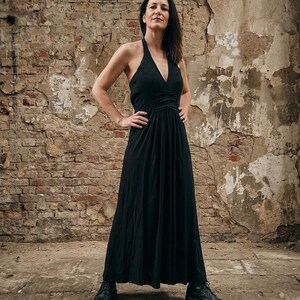 Puede incluir: Un vestido largo negro tipo halter con un escote en V profundo, un corpiño ajustado con cintura fruncida y una falda fluida. La prenda se combina con botines negros con cordones, creando un conjunto uniforme en una paleta de colores oscuros.