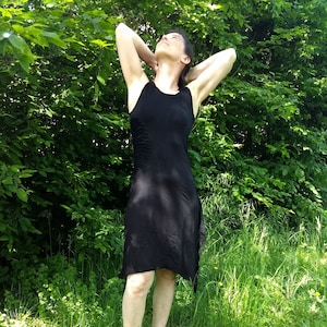 Puede incluir: Una mujer con un vestido negro sin mangas con un detalle fruncido en el lateral, de pie en una zona cubierta de hierba con árboles verdes en el fondo.