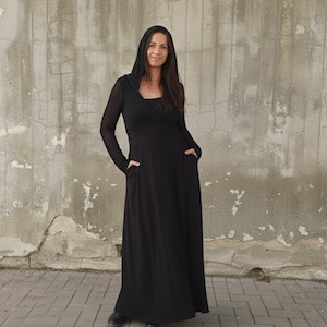 May include: A long black hooded dress with long sleeves and pockets. The dress is made of a soft fabric and has a simple, elegant design. The model is wearing black shoes.