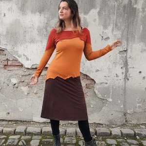 Può includere: Un completo a due pezzi con un top a maniche lunghe e una gonna. Il top sfuma dal rosso all'arancione, con maniche lunghe. La gonna è marrone tinta unita. L'outfit è abbinato a leggings e stivali neri.